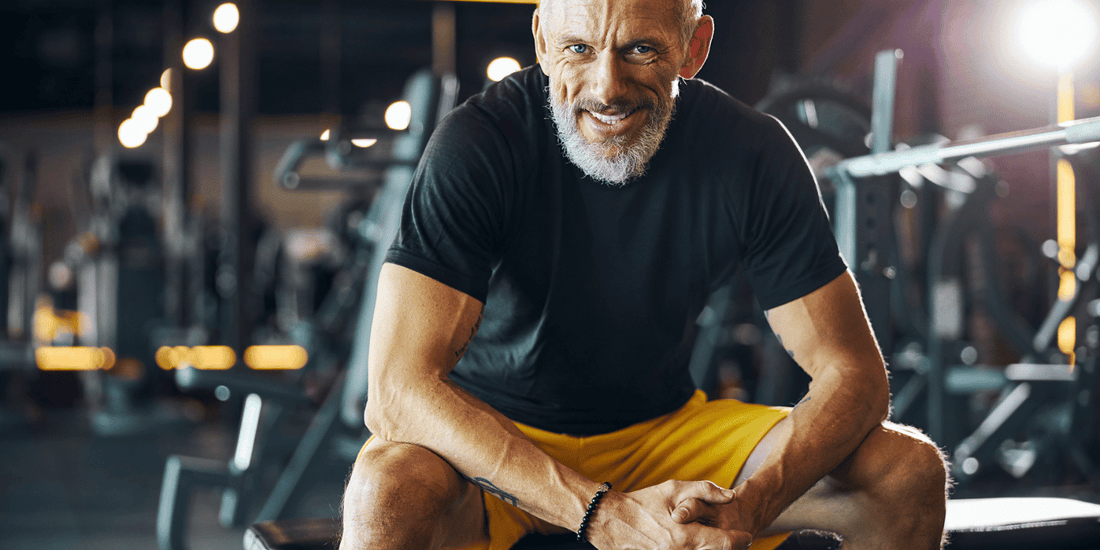  An older man sitting on a gym bench, looking relaxed and observing the workout area around him.
