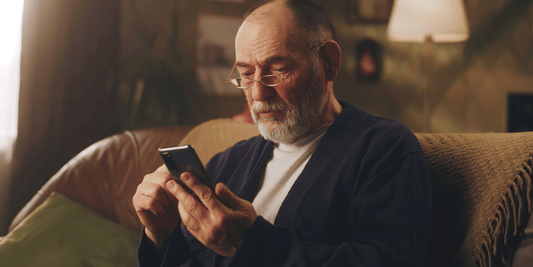 An older man sits on a couch, focused on his phone, with a thoughtful expression on his face.