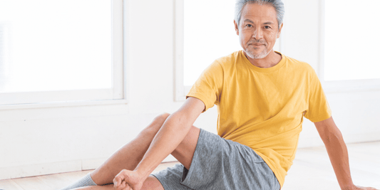 An older man sits on the floor, stretching his leg with a focused expression on his face.