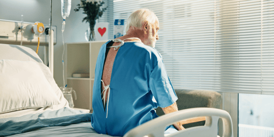 An elderly man in a hospital gown is seated on a hospital bed, with various medical equipment visible around him.