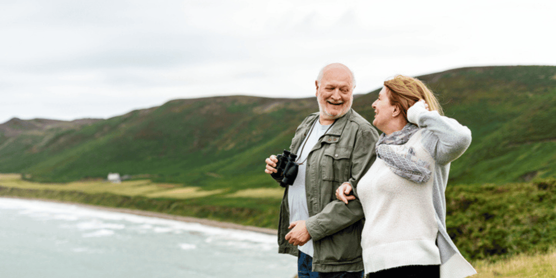 A senior couple stands together on a hill, admiring the expansive ocean landscape.