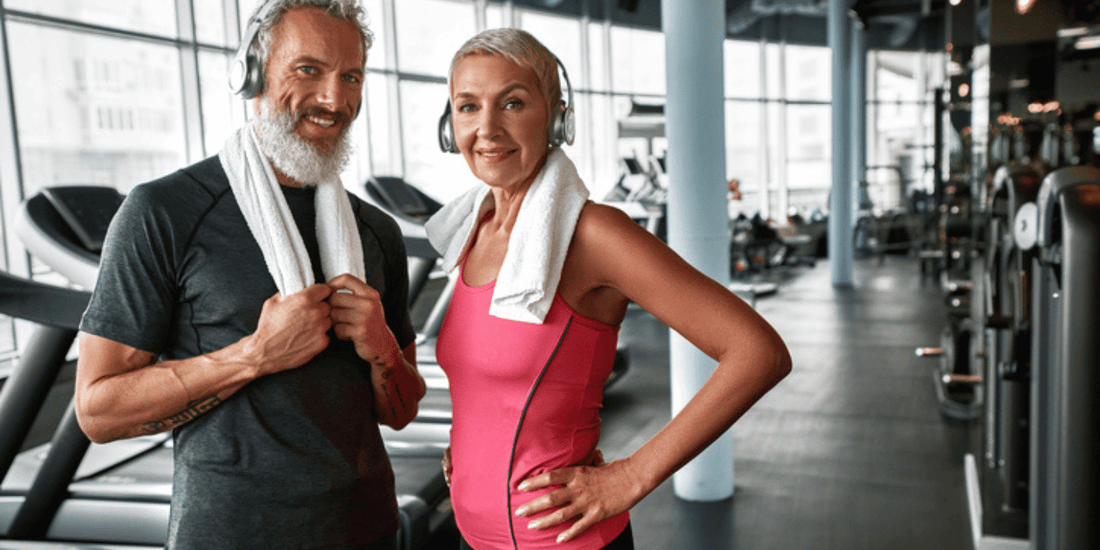 A senior couple poses in a gym, both wearing athletic clothing, with various fitness machines visible in the background.