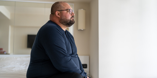 Middle-aged man sitting thoughtfully on the edge of a bed, illustrating difficulty maintaining an erection and the need for erectile dysfunction support.