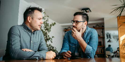 Two middle-aged men having a thoughtful conversation over coffee, illustrating discussion and decision-making when comparing medical penile pump options for erectile dysfunction.