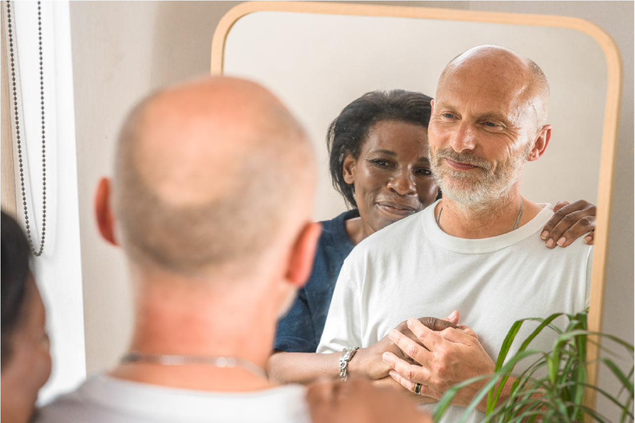 Man looking at a mirror while women stands hugging him from behin