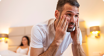 Man at edge of bed holding face with a women in background